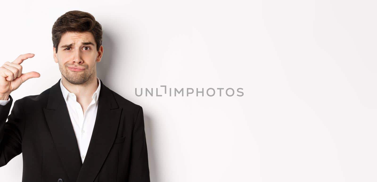 Close-up of handsome businessman in trendy suit, showing something small and looking disappointed, standing against white background.