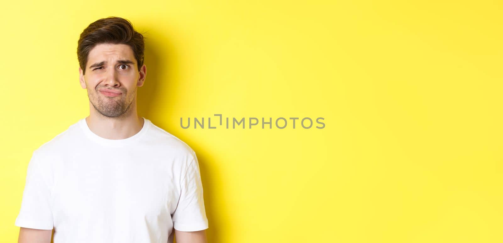Close-up of dispelased young man in white t-shirt looking doubtful, grimacing unsatisfied, standing over yellow background.