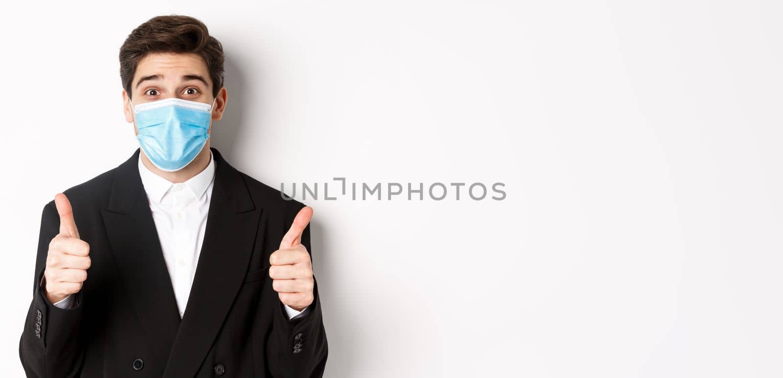 Concept of covid-19, business and social distancing. Close-up of happy businessman in black suit and medical mask, showing thumbs-up, making a compliment, white background.