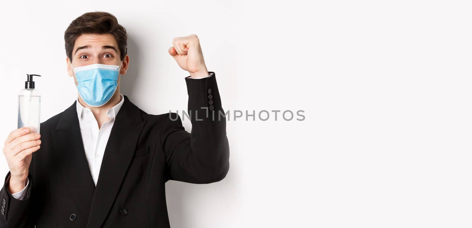 Concept of covid-19, business and social distancing. Close-up of happy man in trendy suit and medical mask, cheering and raising hand up, showing hand sanitizer, white background.