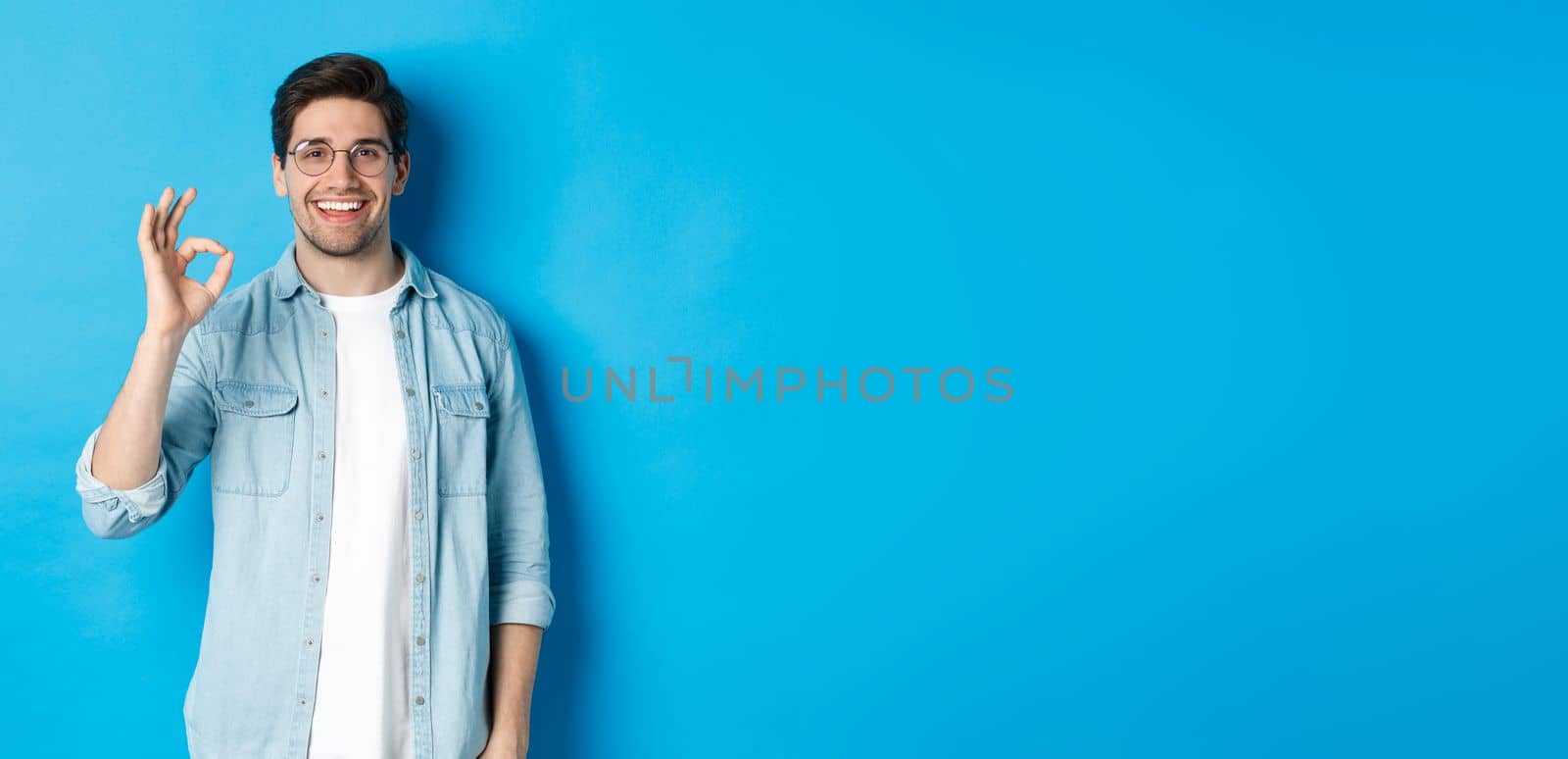 Attractive young man wearing glasses and casual clothes, showing ok good sign in approval, like something, standing against blue background.