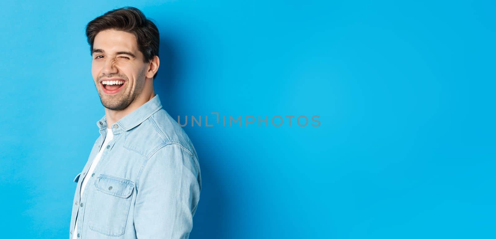 Happy and confident man turn head at camera, winking and smiling, standing over blue background.