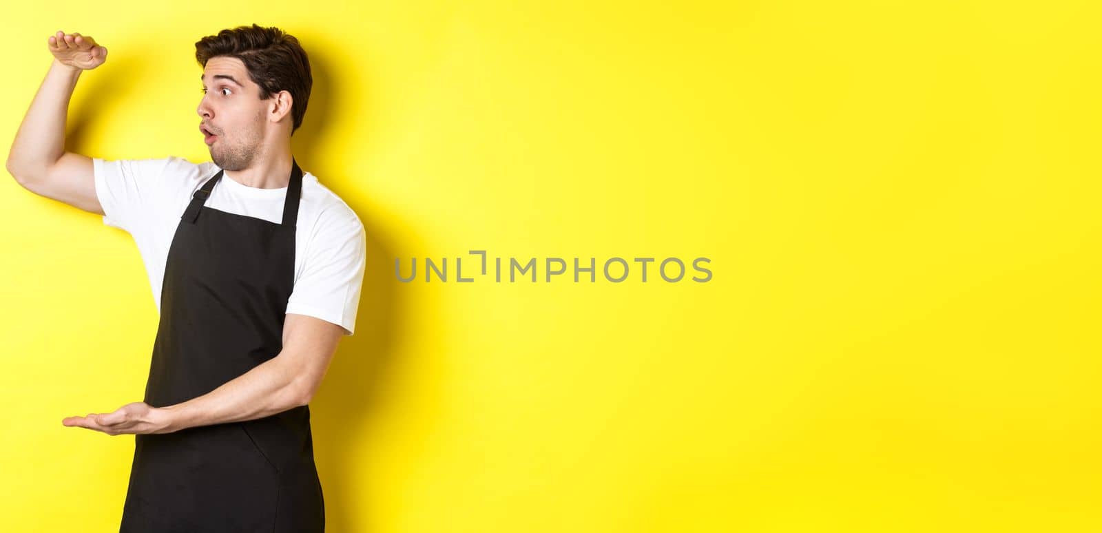 Waiter in black apron looking amazed at something large, holding big object, standing over yellow background.