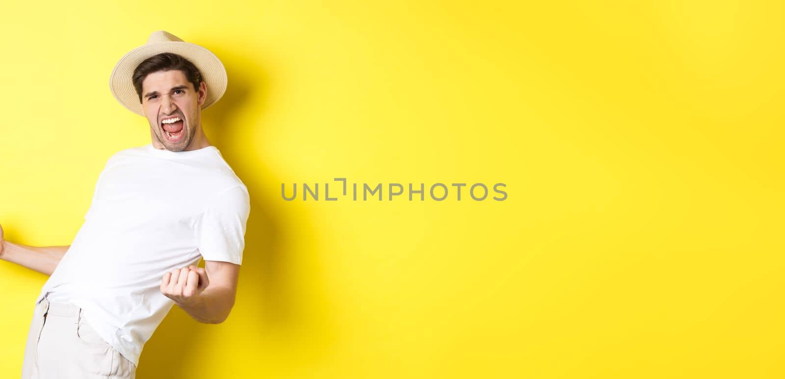 Concept of tourism and vacation. Satisfied young male tourist celebrating, winning something and rejoicing, making fist pump and shouting yes, standing against yellow background.