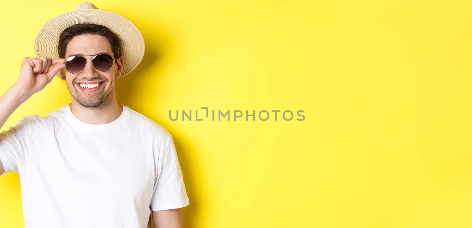 Concept of tourism and vacation. Close-up of handsome man tourist looking happy, wearing sunglasses and summer hat, standing over yellow background by Benzoix