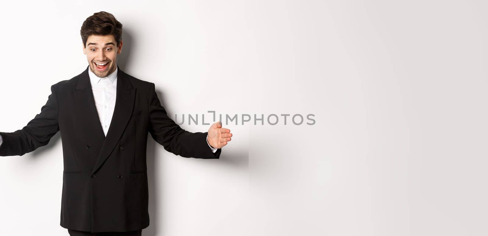 Portrait of excited handsome man in suit, shaping big object on copy space and smiling amazed, holding something, standing over white background.