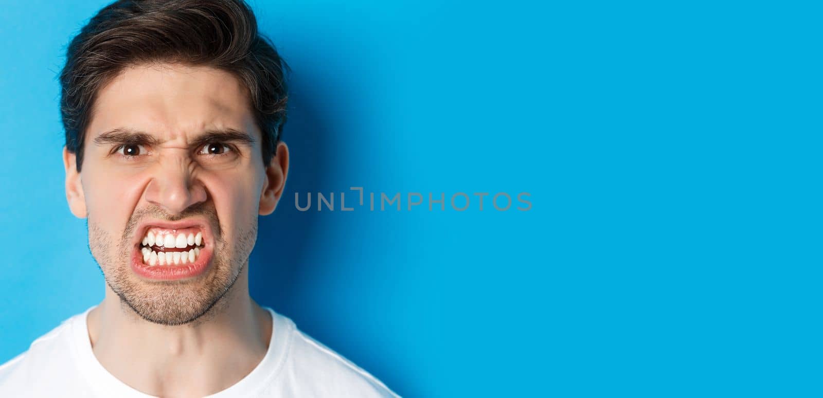 Headshot of angry caucasian man looking with scorn and dismay, being mad at person, standing against blue background.