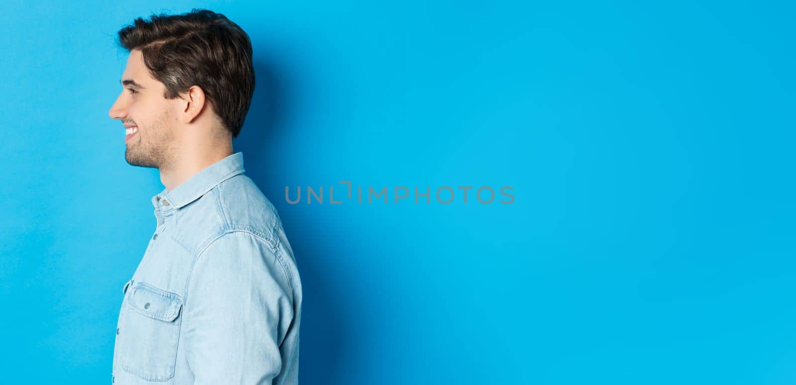 Profile of handsome young man looking left, smiling happy, standing over blue background.