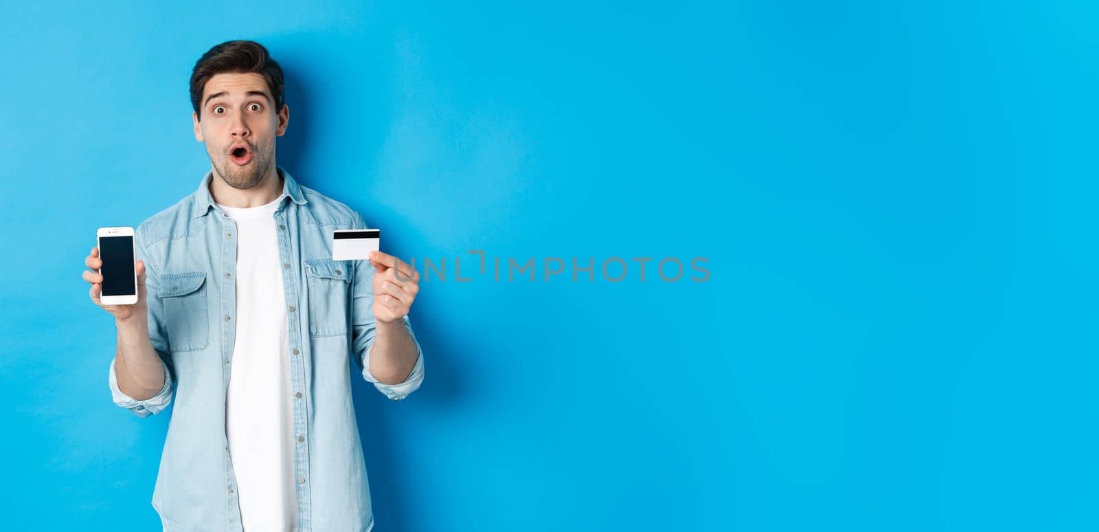 Amazed young man showing mobile cell phone screen and credit card, shop online, standing against blue background.