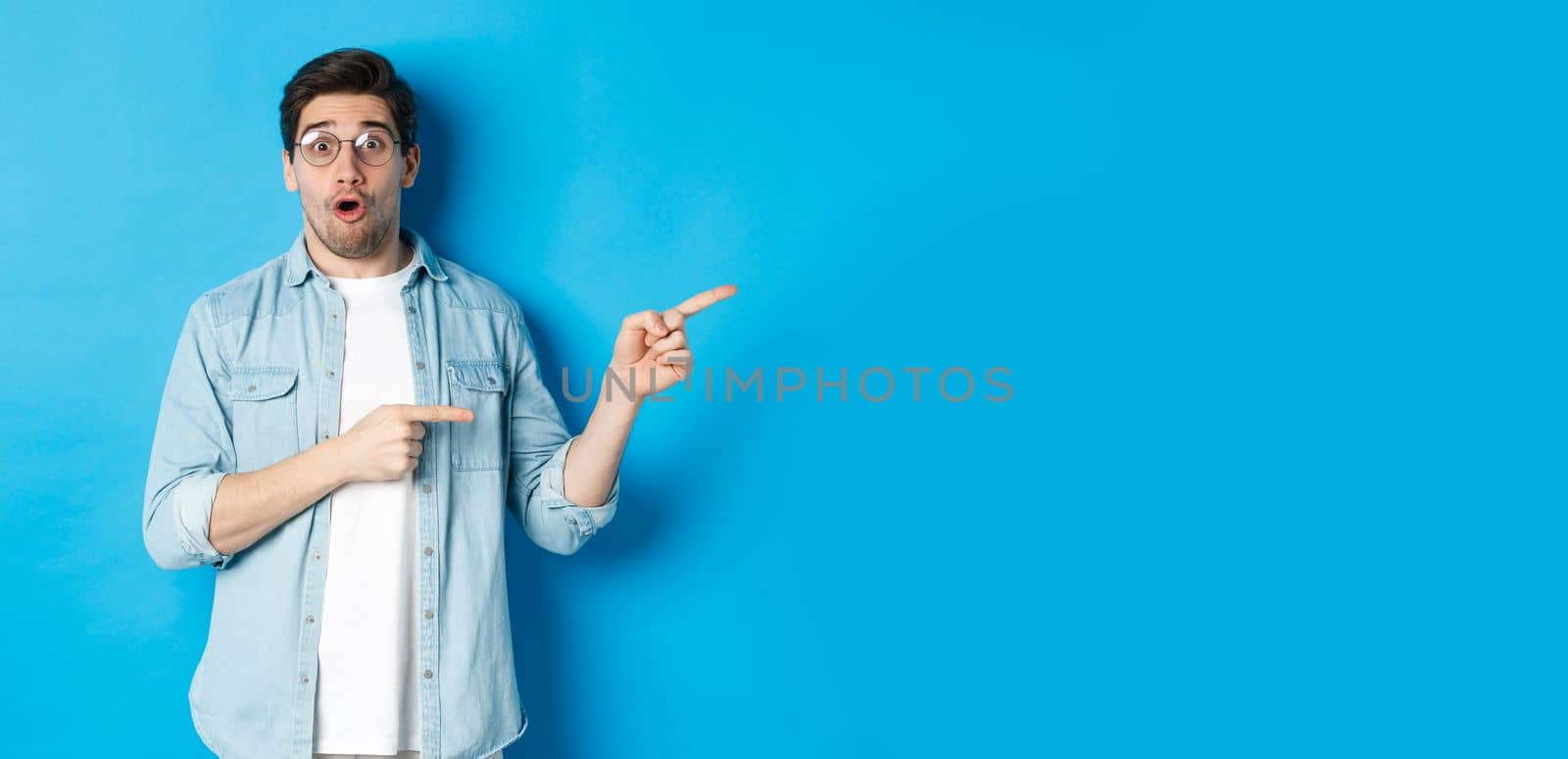 Surprised man in glasses pointing right at copy space, showing promo offer on blue background, standing over blue background by Benzoix