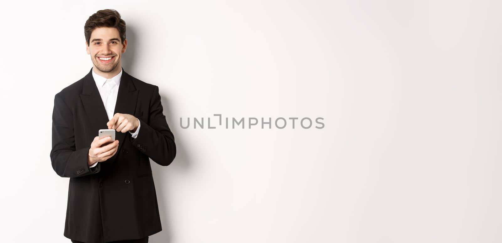 Image of handsome businessman in suit showing you app on smartphone, pointing finger at mobile screen and smiling, standing over white background.