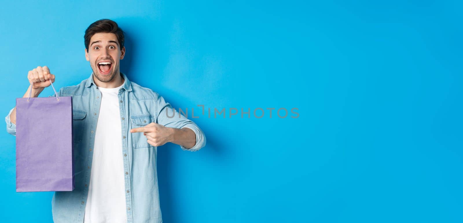 Concept of shopping, holidays and lifestyle. Excited guy pointing finger at paper bag and looking amazed, recommending store, announcing discounts, blue background.