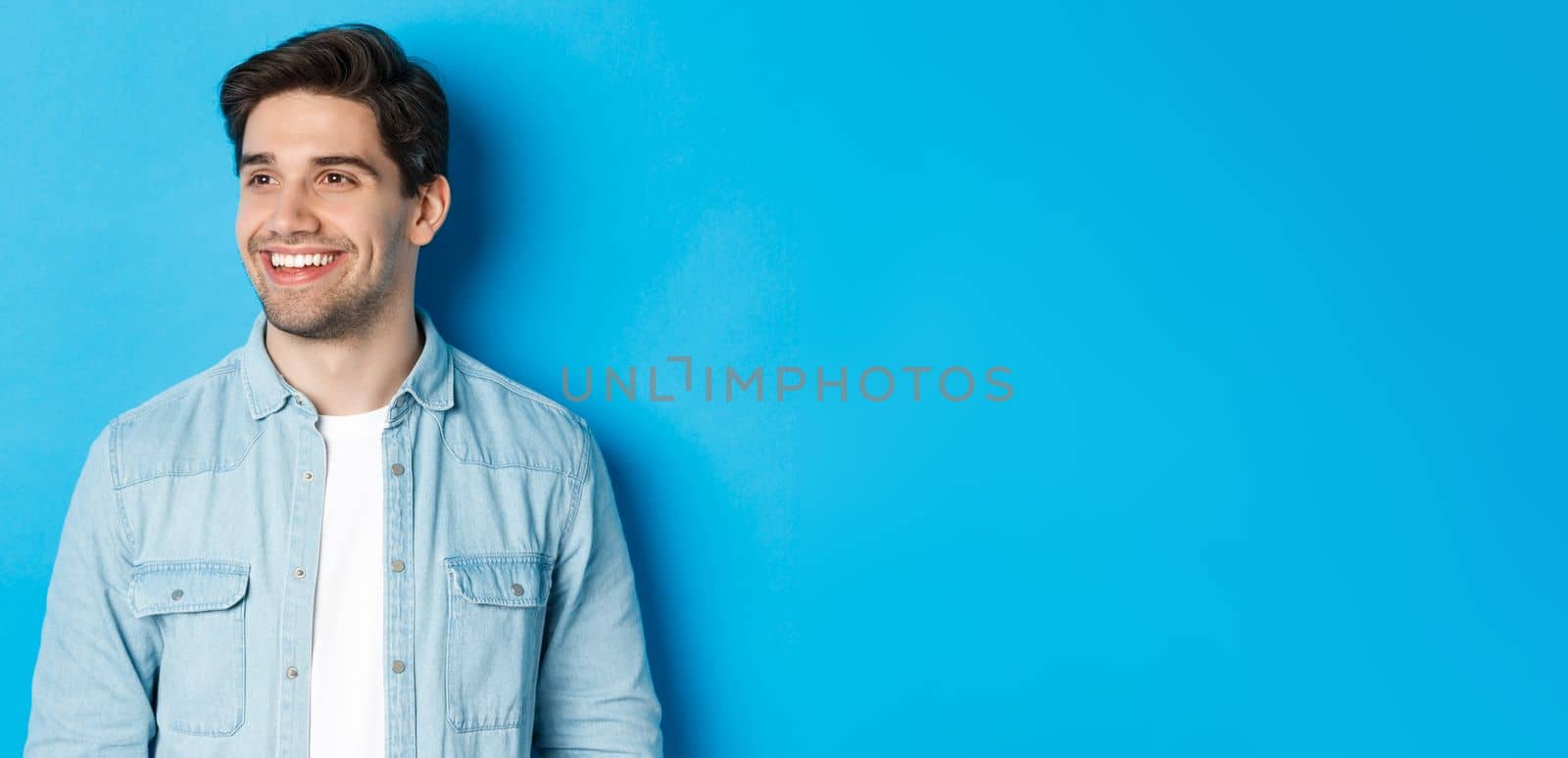 Young modern guy with beard, looking left and smiling pleased at copy space, standing over blue background.