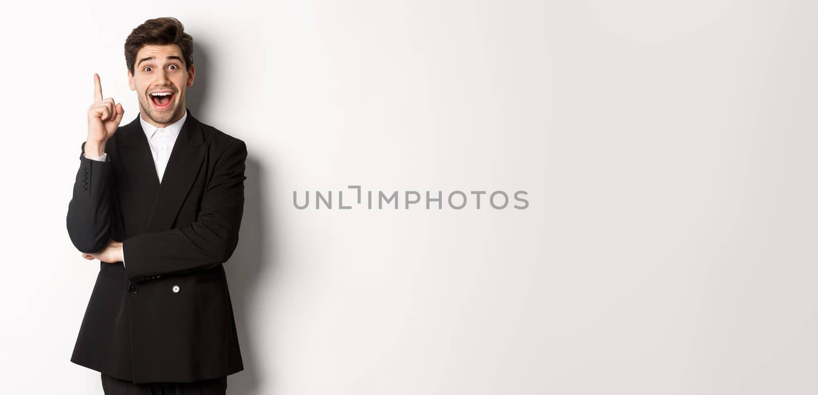 Portrait of attractive man in suit, having an idea, standing excited and raising one finger to tell suggestion, think-up solution, standing over white background.