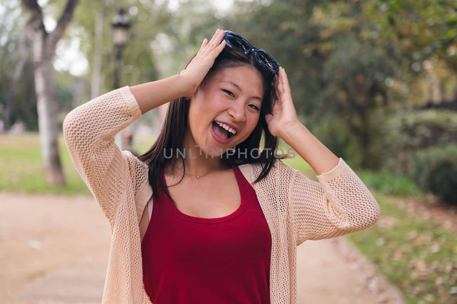 cheerful asian woman puts on her sunglasses on her hair laughing in the park, concept of youth and beauty