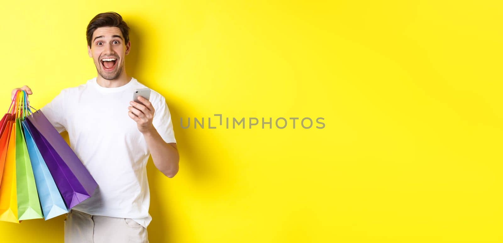 Image of happy man receive cashback for purchase, holding smartphone and shopping bags, smiling excited, standing over yellow background.