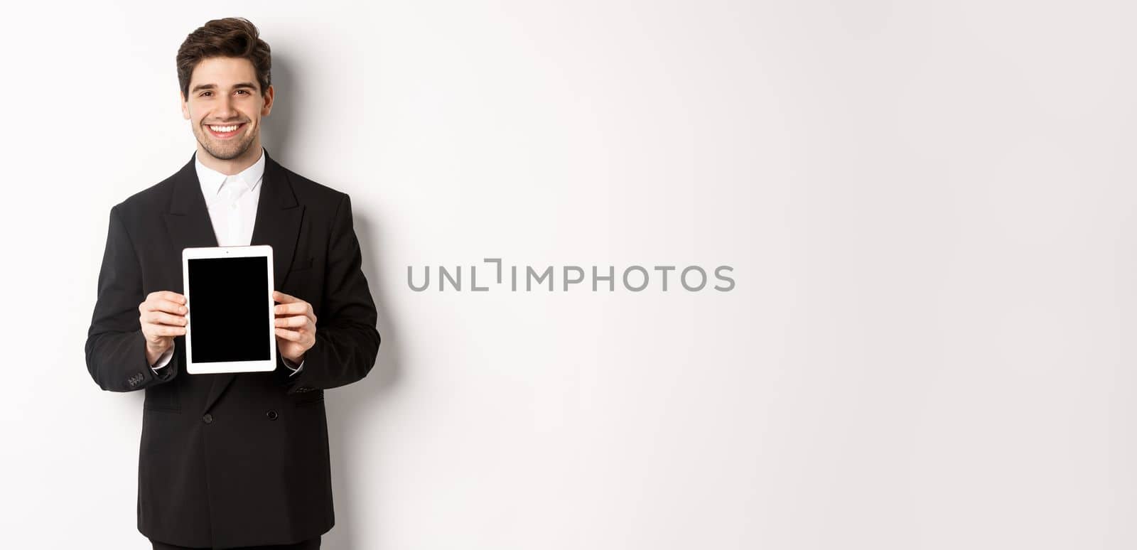 Portrait of handsome businessman in trendy suit, showing digital tablet screen and smiling, standing against white background.