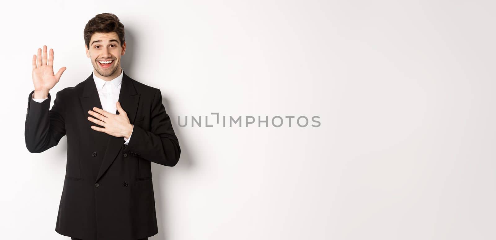 Image of handsome smiling guy telling the truth, raising on arm to promise something, standing over white background in black suit.