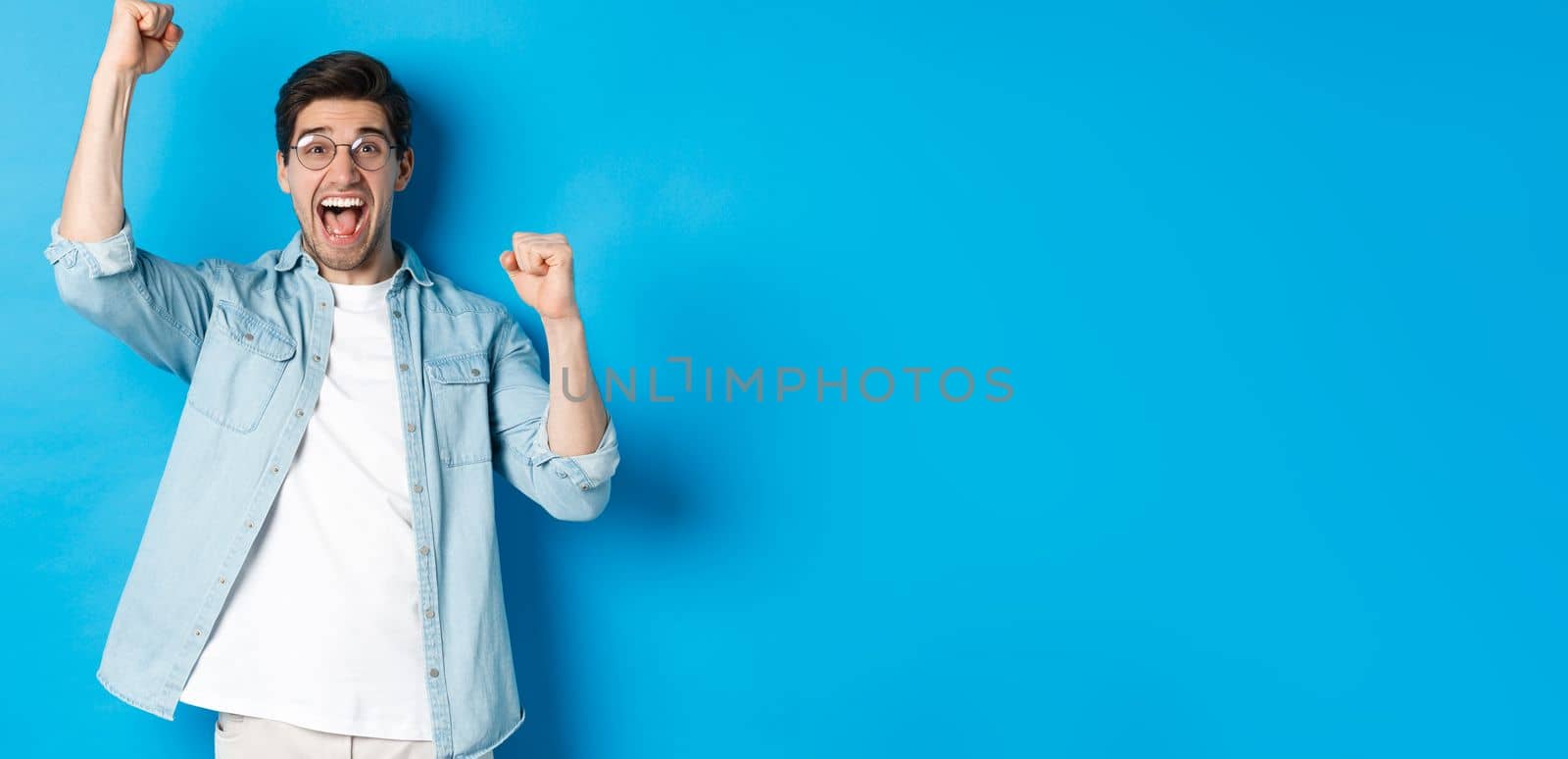 Excited handsome man triumphing, raising hands up and shouting for joy, celebrating victory, standing against blue background.