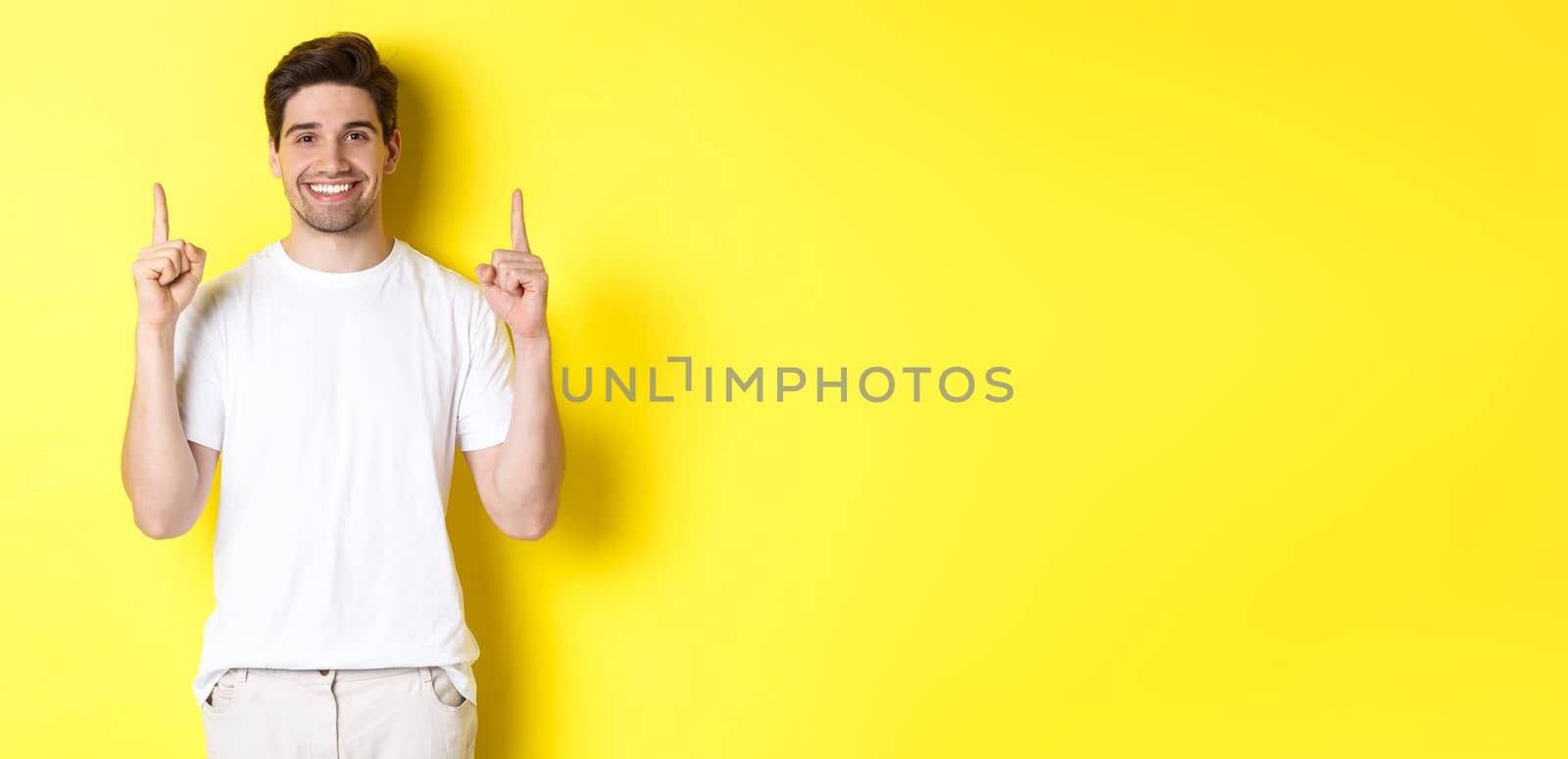 Handsome guy in white t-shirt pointing fingers up, showing shopping offers, standing over yellow background.