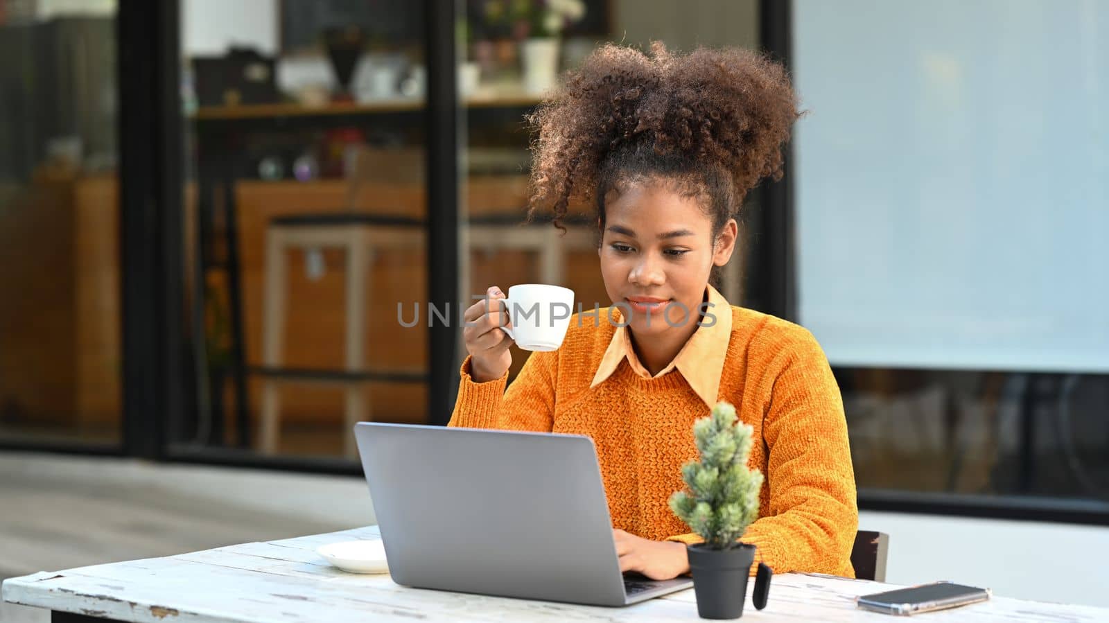 Pleased female freelancer drinking coffee and working online on laptop while sitting outdoor.