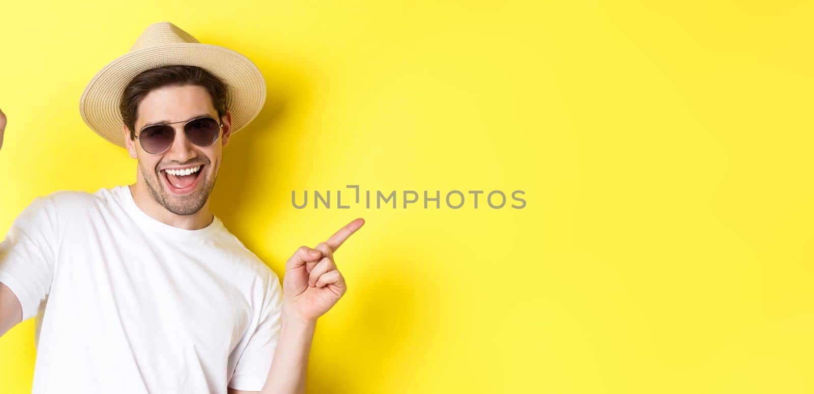 Concept of tourism and vacation. Close-up of man enjoying holidays on trip, dancing and pointing fingers sideways, wearing sunglasses with straw hat.