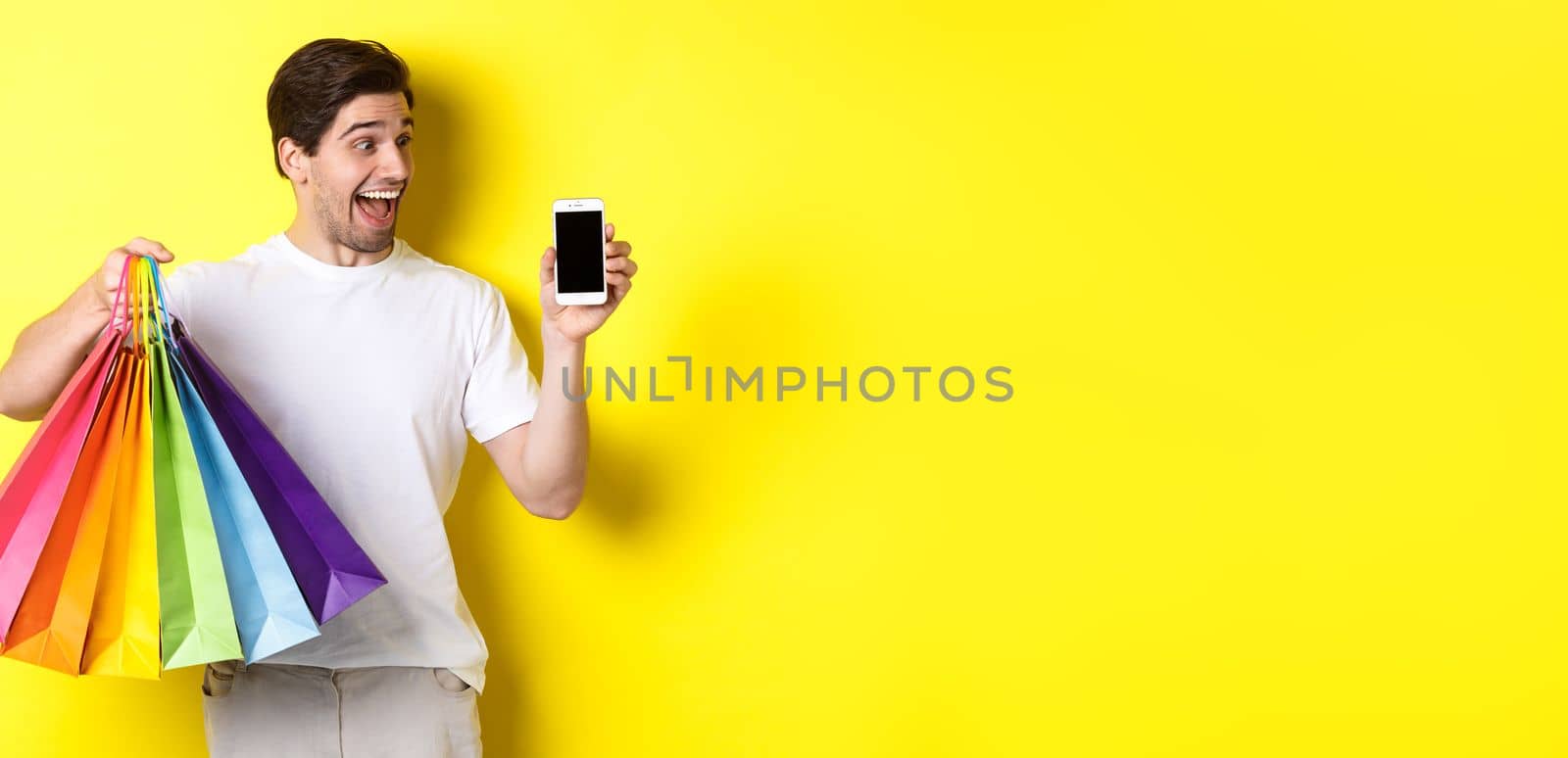 Young man holding shopping bags and showing mobile phone screen, money application, standing over yellow background.