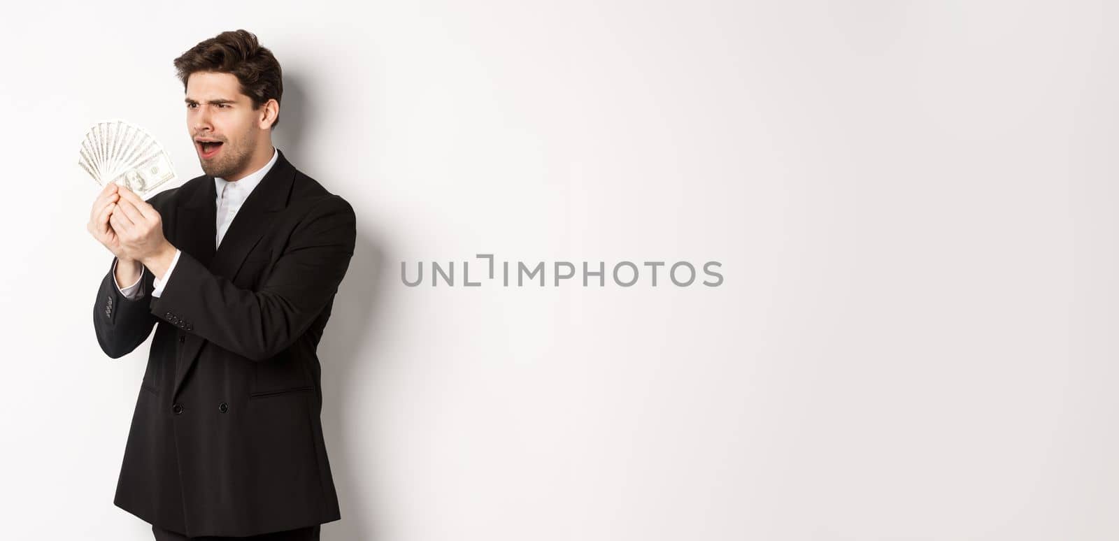 Image of confused businessman looking at fake money, standing over white background in black suit.