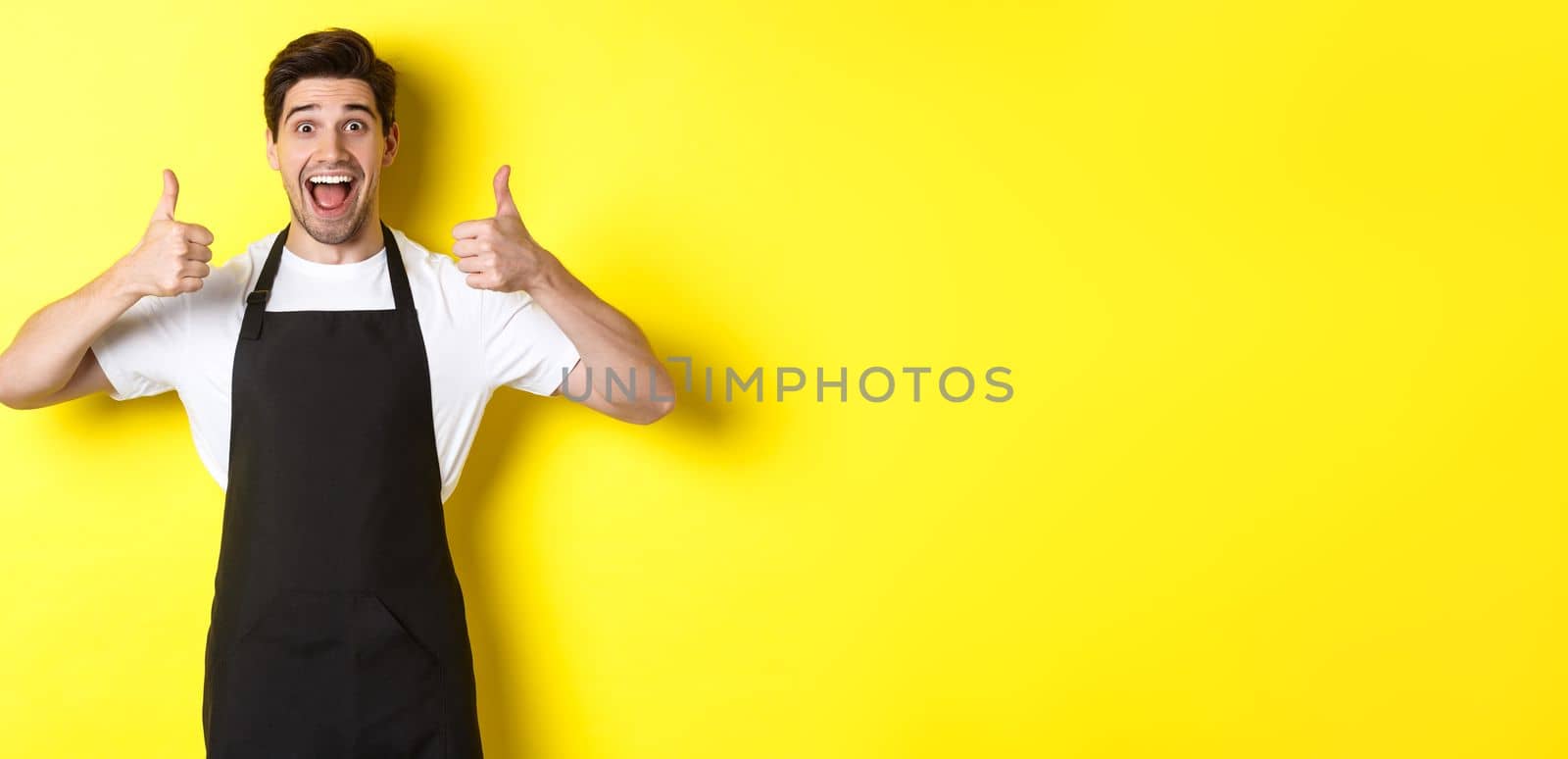 Cheerful man barista in black apron showing thumbs-up, recommending cafe or restaurant, standing against yellow background.