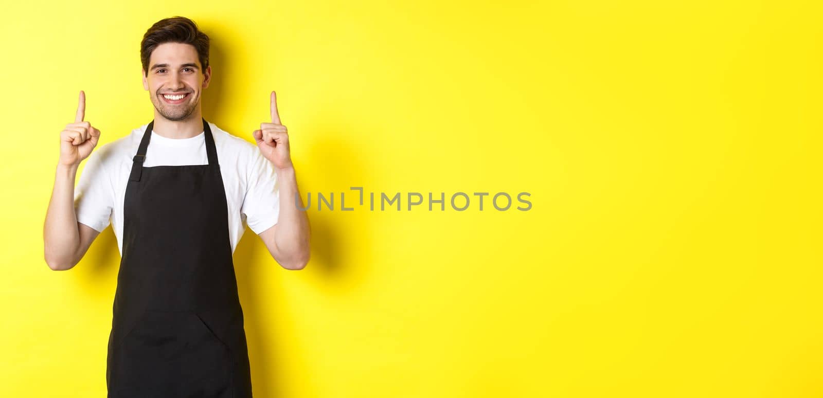 Handsome barista showing advertisement, coffee shop promo offer, pointing fingers up, standing against yellow background.
