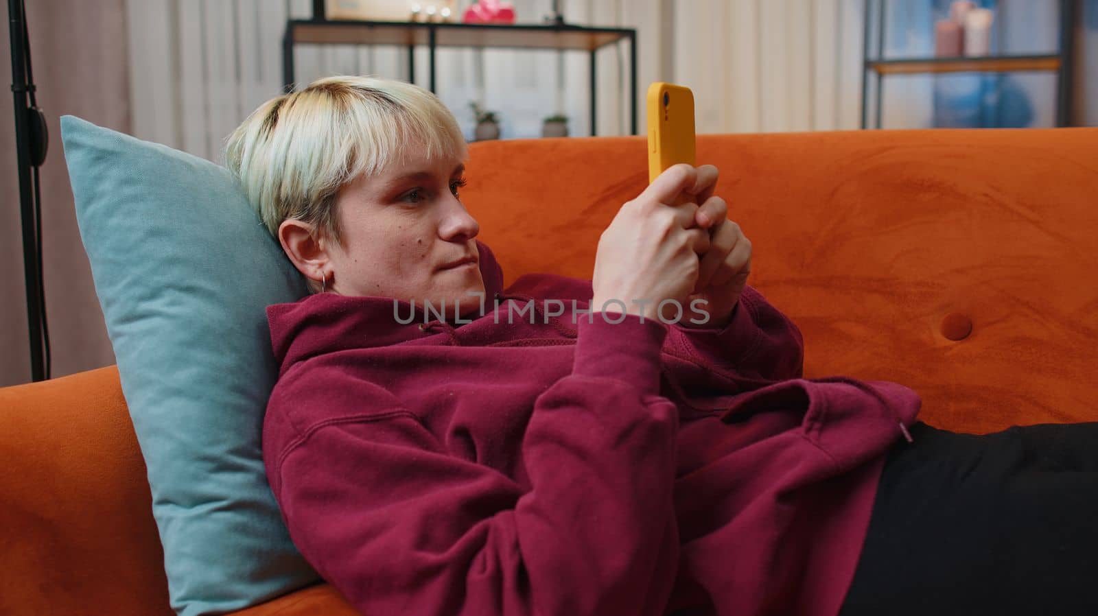 Young woman with short blonde hair lying on sofa uses mobile phone smiles at night evening home. Girl texting share messages content on smartphone social media applications online watching relax movie