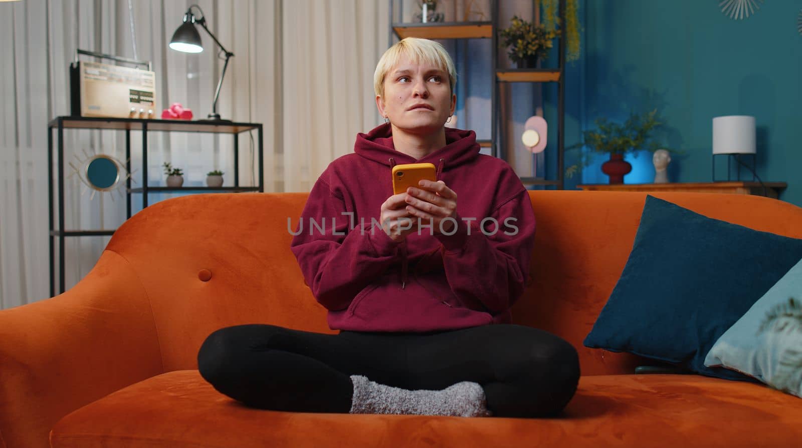 Woman with short blonde hair sitting on sofa uses mobile phone smiles at night home apartment. Girl texting share messages content on smartphone social media applications online, watching relax movie