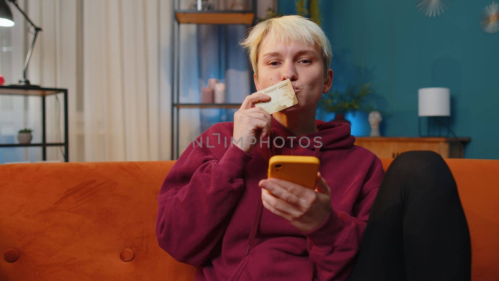 Young woman with short hair using credit bank card and smartphone while transferring money, purchases online shopping, order food delivery at home apartment indoors. Girl in evening room on couch