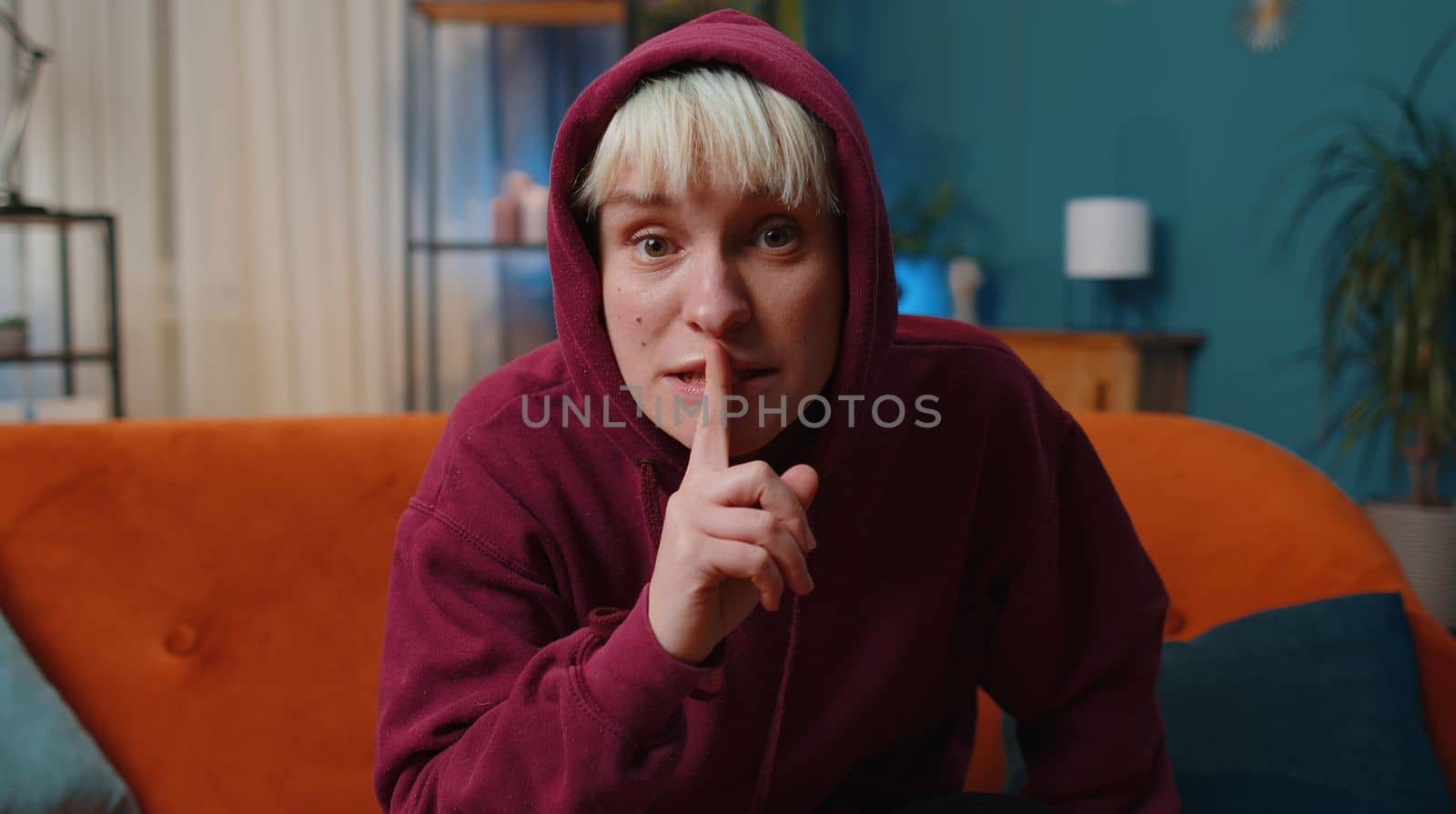 Shh be quiet, hush please. Young woman with short blonde hair presses index finger to lips makes silence hush gesture sign do not tells gossip secret. Girl at modern home evening room sitting on couch