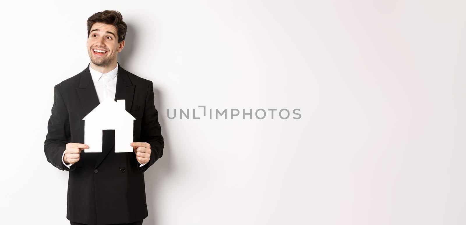 Image of handsome investor in black suit, holding house maket and looking at upper left corner thoughtful, standing against white background.