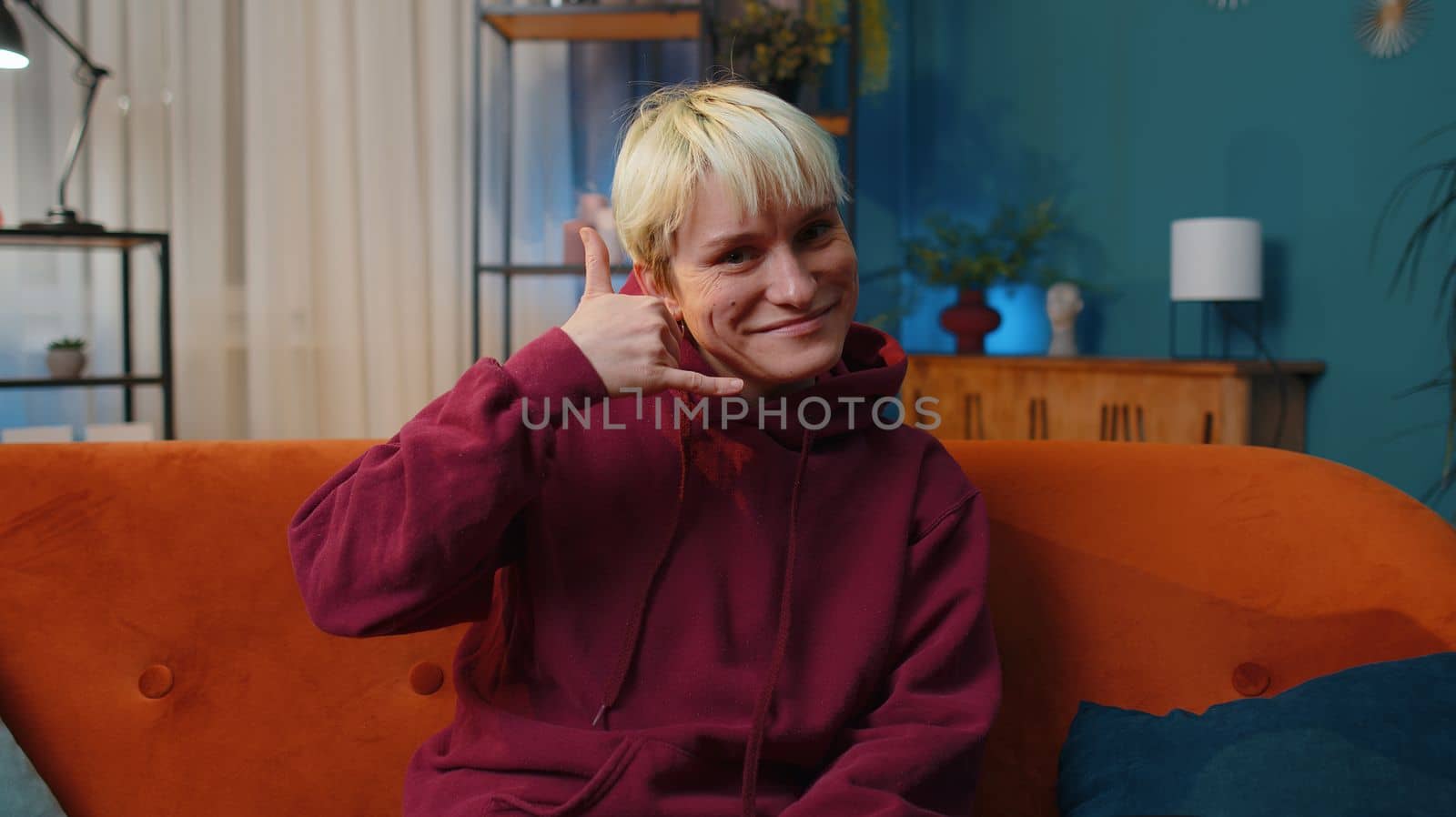 Call me, here is contact number. Young woman with short blonde hair looking at camera advertising doing phone gesture like says hey you call me back at home. Lovely girl at evening room apartment