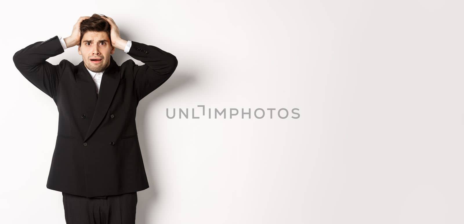 Frustrated and worried businessman in black suit, panicking as looking at trouble, holding hands on head alarmed, standing against white background.