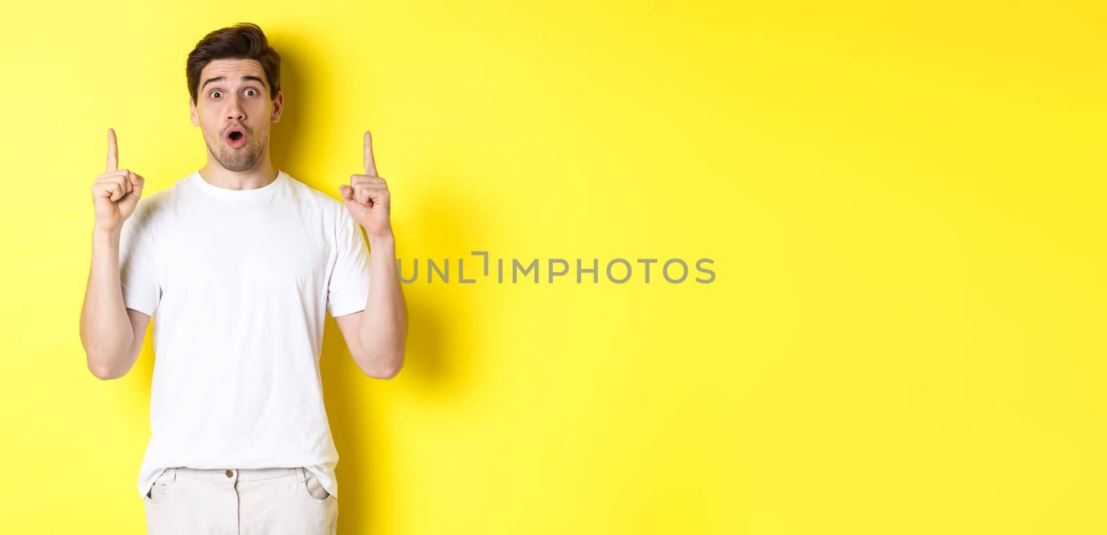 Surprised handsome guy in white t-shirt, pointing fingers up, interested about advertisement, standing against yellow background.