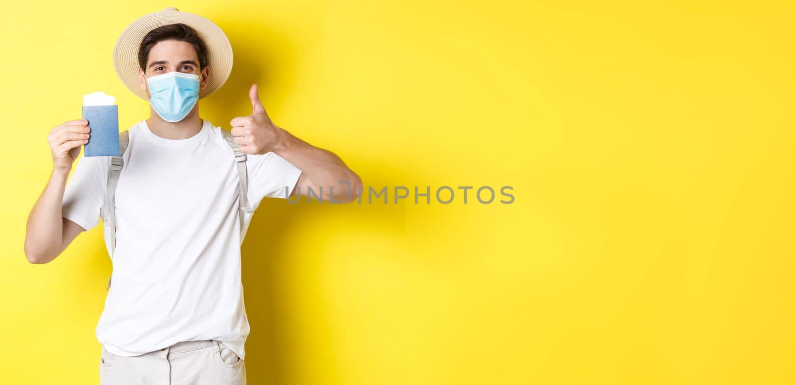 Concept of covid-19, tourism and pandemic. Happy male tourist in medical mask showing passport, going on vacation during coronavirus, make thumb up sign, yellow background.
