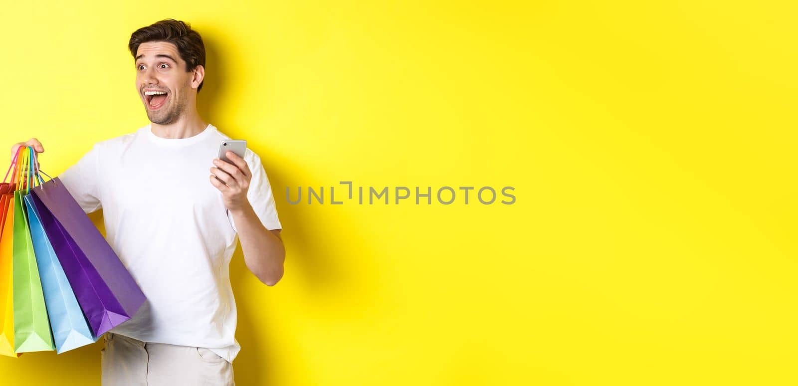 Concept of mobile banking and cashback. Happy man looking amazed, holding shopping bags and smartphone, yellow background by Benzoix