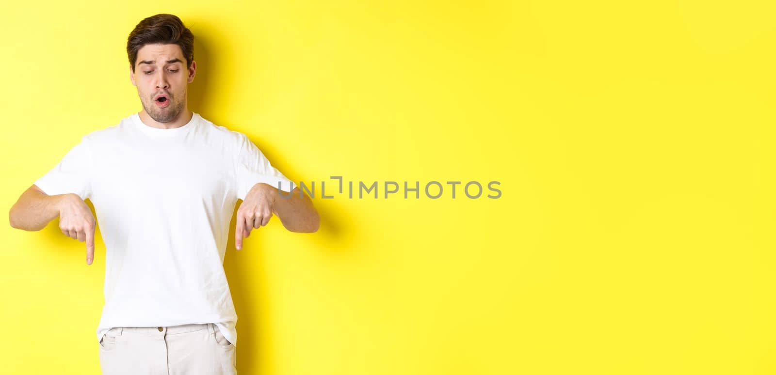 Worried guy pointing fingers and looking down, standing startled against yellow background.