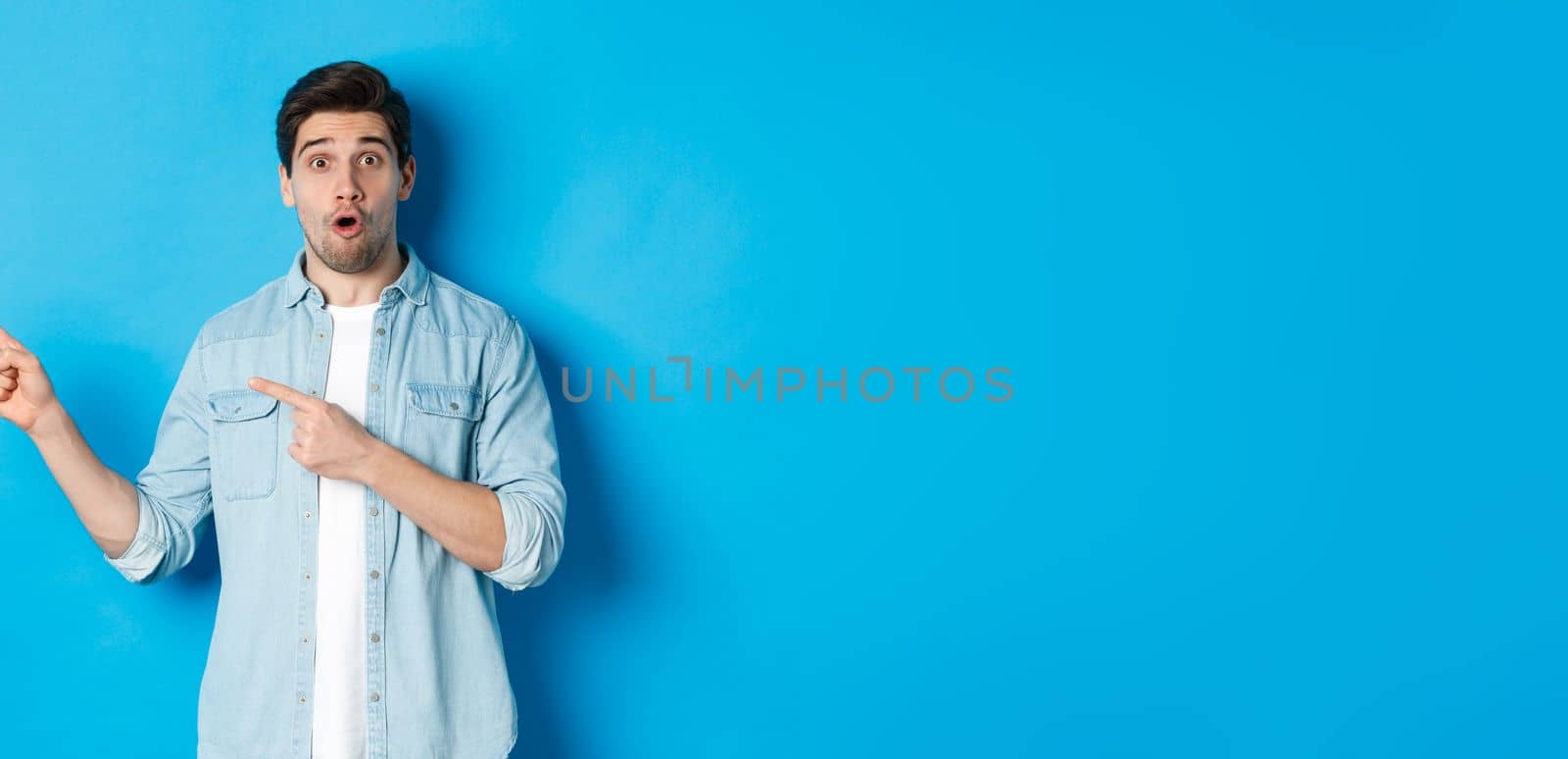 Portrait of surprised adult man in casual clothes showing announcement, pointing fingers left and looking amazed, standing against blue background by Benzoix