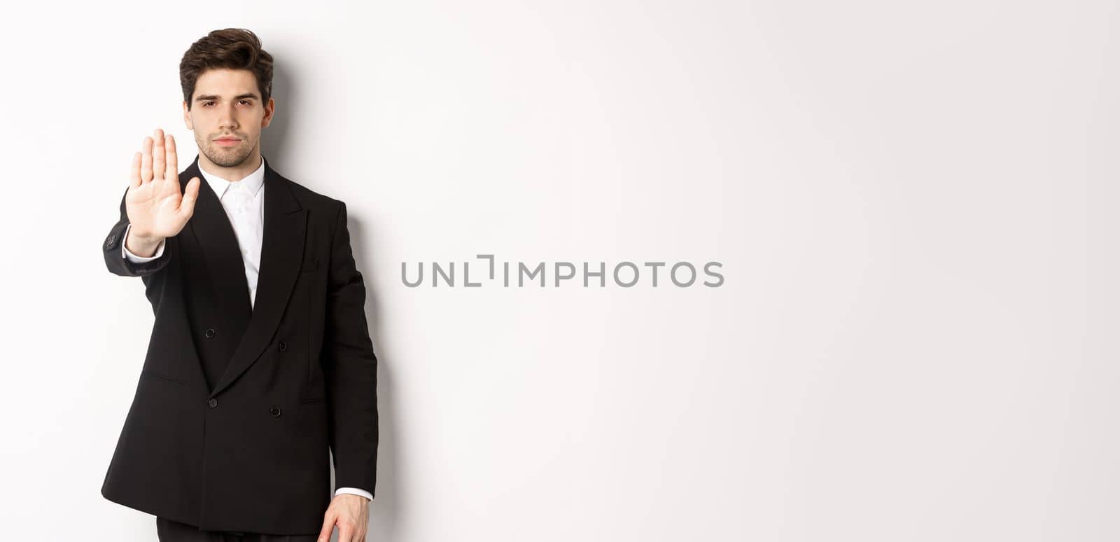 Portrait of serious handsome man in formal suit, extending hand to stop you, prohibit action, forbid and disagree with something, standing against white background.