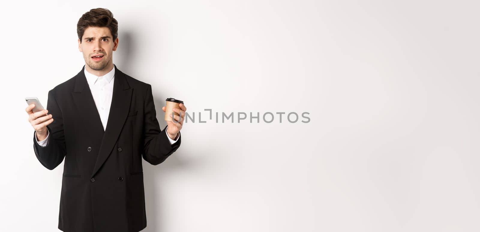 Image of confused businessman standing in a suit, holding cup of coffee and mobile phone, cant understand something, standing against white background by Benzoix