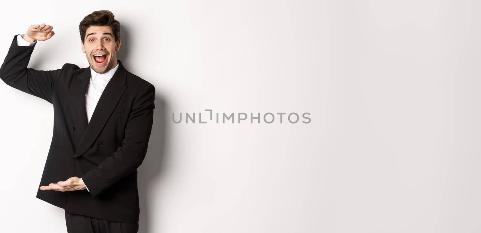 Portrait of excited handsome man in suit, shaping big object on copy space and smiling amazed, holding something, standing over white background by Benzoix