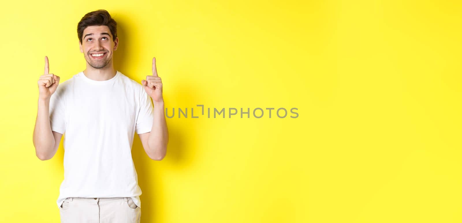 Excited attractive man in white t-shirt, pointing fingers up, looking at advertisement with happy smile, standing over yellow background.