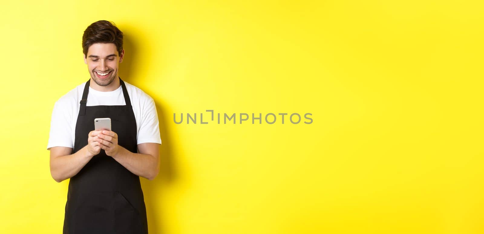 Waiter in black apron reading message on mobile phone, smiling happy, standing over yellow background.