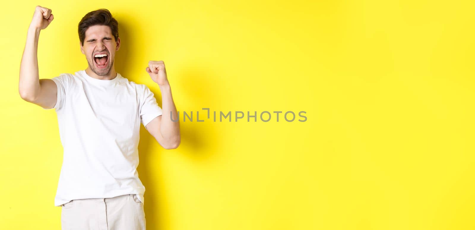 Excited handsome man winning and shouting for joy, raising hand up and triumping, achieve goal, standing over yellow background.