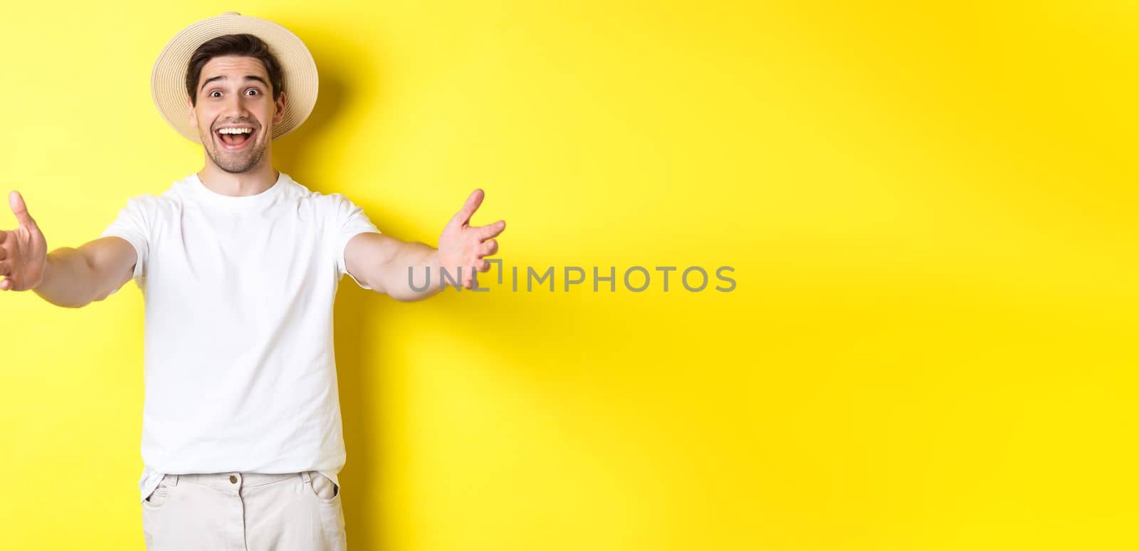 Concept of tourism and summer. Happy and friendly man traveller reaching hands for hug, greeting or welcome you, standing over yellow background by Benzoix