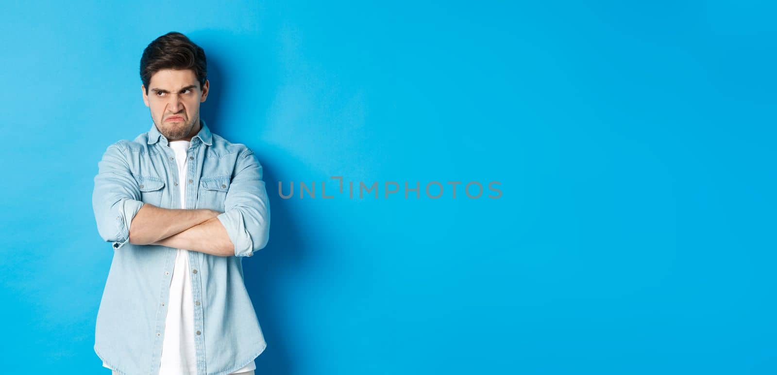 Angry guy cross arms on chest and looking away with insulted expression, standing offended against blue background.